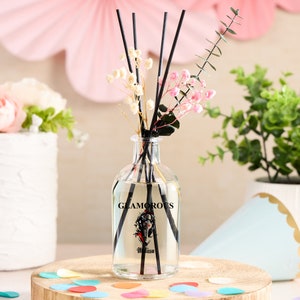 May include: A clear glass diffuser bottle with black reeds, dried flowers, and the word "GLAMOROUS" printed on the front. The bottle contains a clear liquid and is sitting on a wooden slice. Decorative confetti is scattered around the base.