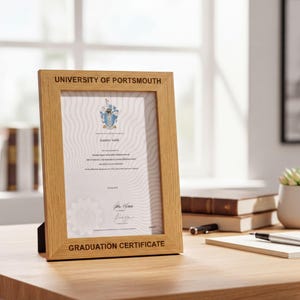 A wooden frame containing a graduation certificate sits on a wooden desk, with a stack of books and a pen nearby.