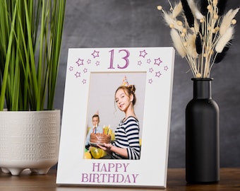 May include: White photo frame with a pink 13 and stars around the border. The frame says "Happy Birthday" at the bottom. A photo of a girl holding a birthday cake is in the frame.