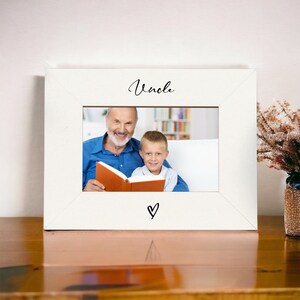 以下が含まれることがあります： 白い写真フレームに、年配の男性と若い男の子が一緒に本を読んでいる写真が飾られています。フレームには「おじ」という言葉が書かれており、写真の真下にハートが描かれています。
