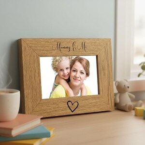 A wooden picture frame with a photo of a woman and a child, accompanied by a teddy bear and a stack of books, sits on a desk near a window.