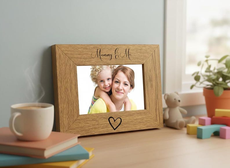 A wooden picture frame with a photo of a woman and a child, accompanied by a teddy bear and a stack of books, sits on a desk near a window.