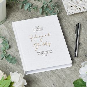 May include: White wedding planner with gold lettering that says "Our Wedding Planner Hannah & Gibby Things to do before we say I do."