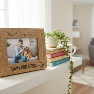 A wooden picture frame with a photo of a man and a dog, along with the text "Best day dad" and "Adam and Bruno" engraved on it, sits on a shelf in a living room.