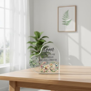 May include: A clear acrylic heart-shaped ornament with the word "Gran" in script. The ornament features floral and butterfly accents, with the text "Wishing you a very Happy Birthday, Love Jesse & Sandra XX."