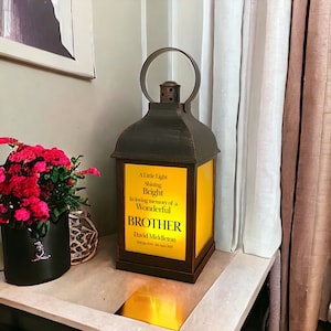 May include: A black metal lantern with a yellow glass panel. The glass panel has a message that reads "A Little Light Shining Bright In loving memory of a Wonderful BROTHER David Middleton 20th Jan 1941 - 04th April 2023"