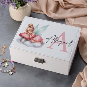 May include: White jewellery box with a pink and green fairy illustration, a pink letter A, and the name Abigail. The box has a silver latch.