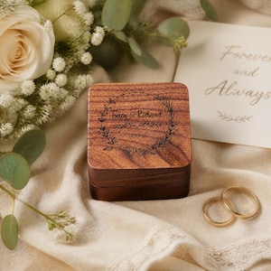 A wooden box with a wreath design, two gold wedding rings, and a note that says "Forever and Always" are arranged on a white cloth.
