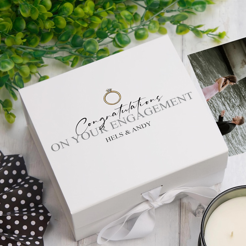 A white gift box with a gold ring on it, placed on a white surface next to a candle and some greenery.