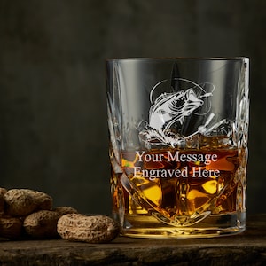 May include: A clear glass whiskey tumbler with a cut-glass design. The glass is filled with amber liquid and features an etched design of a fish jumping out of the water. The text "Your Message Engraved Here" is etched on the glass.