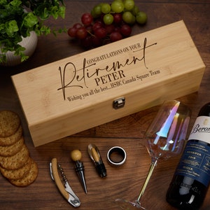May include: A wooden box with a hinged lid, engraved with the text "Congratulations on your Retirement Peter Wishing you all the best...HSBC Canada Square Team". The box contains a wine bottle opener, a bottle stopper, a wine pourer, and a bottle stopper ring. The box is sitting on a wooden surface with a wine glass and a bottle of wine.