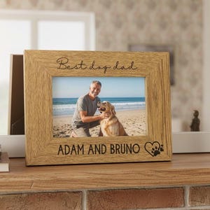 A wooden picture frame with a photo of a man and a dog, along with the text "Best day dad" and "Adam and Bruno" engraved on it. The frame is placed on a wooden shelf, and there is a potted plant and some figurines nearby.