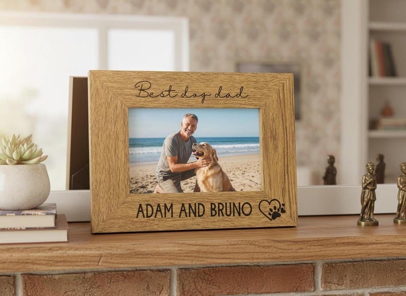 A wooden picture frame with a photo of a man and a dog, along with the text "Best day dad" and "Adam and Bruno" engraved on it. The frame is placed on a wooden shelf, and there is a potted plant and some figurines nearby.