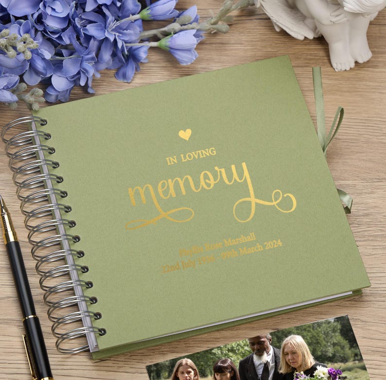May include: A green spiral bound memory book with gold foil lettering that reads "In loving memory" and a small heart. The book is personalized with the name "Marie Rose Marshall" and the dates "22nd July 1954 - 20th March 2024".