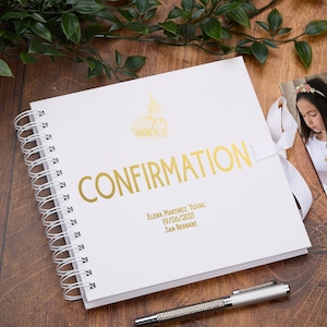 Puede incluir: Un libro de visitas de Confirmación blanco con encuadernación en espiral, letras doradas y una ilustración de una iglesia. El libro está atado con una cinta blanca e incluye el texto "CONFIRMATION" y detalles personalizados. Un bolígrafo plateado descansa sobre la superficie de madera.