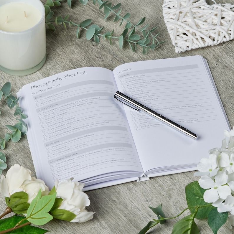 Puede incluir: Un cuaderno blanco de lista de verificaci&oacute;n de fotograf&iacute;a de boda con un bol&iacute;grafo plateado abierto sobre una superficie de madera. El cuaderno tiene una lista de verificaci&oacute;n para fotos de antes de la boda, la ceremonia y la recepci&oacute;n.