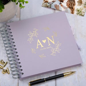 Peut inclure: Un album photo de couleur lavande avec une reliure spirale dorée. La couverture avant présente une couronne florale dorée avec les initiales "A & N" et la date "23.05.2025" au centre.