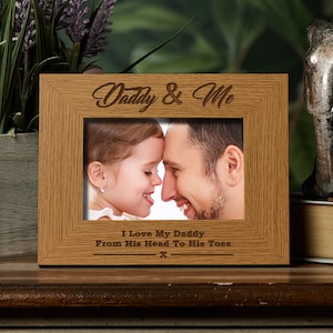 May include: A wooden photo frame with a light brown finish. The frame has an engraved inscription that reads "Daddy & Me" and "I Love My Daddy From His Head To His Toes." The frame includes a photo of a father and daughter.
