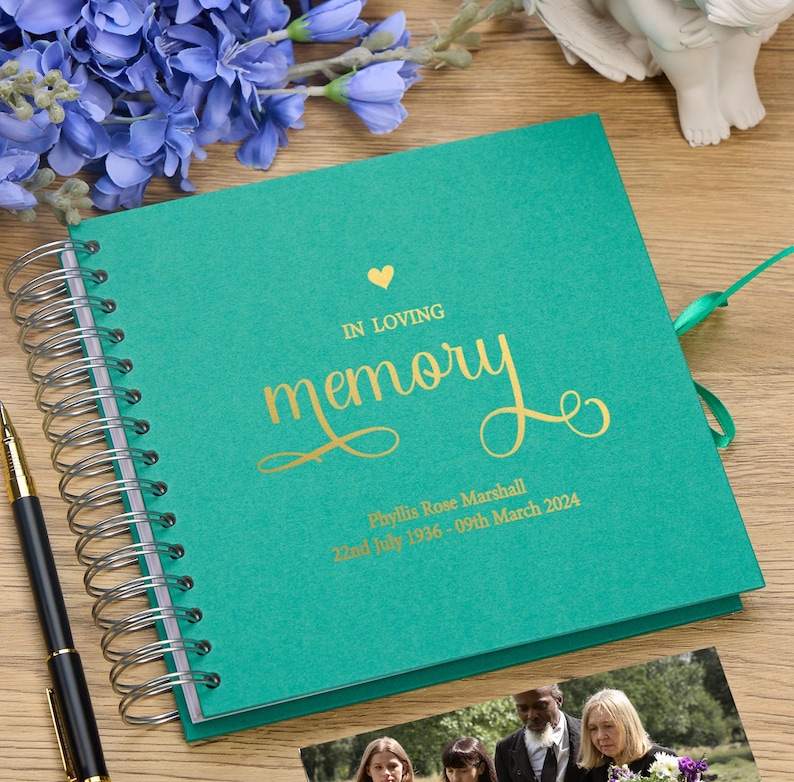 May include: A teal green memory book with a gold heart and the words "In Loving Memory" in gold script. The book has a gold ribbon and a personalized inscription that reads "Phyllis Rose Marshall 22nd July 1936 - 09th March 2024".