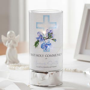 May include: A cylindrical, illuminated candle holder with a silver base. The white surface features a blue cross, blue flowers, and the text "FIRST HOLY COMMUNION" with a name and message. A small angel statue is in the background.