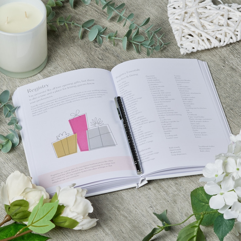 Puede incluir: Libro de registro de bodas blanco abierto con un bol&iacute;grafo en la p&aacute;gina. El libro tiene una secci&oacute;n para regalos y una secci&oacute;n para art&iacute;culos esenciales de cocina y comedor. El libro est&aacute; rodeado de flores blancas y una vela blanca.