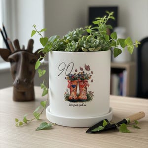 May include: A white ceramic plant pot with a succulent and trailing plant. The pot has a design of wellington boots filled with flowers, the number 90, and the text "Love from Jack xx".