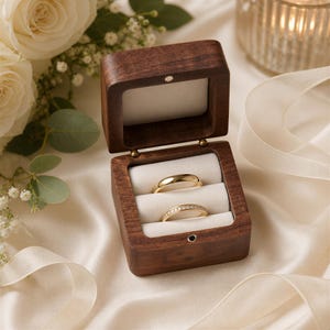 A wooden box containing two gold wedding rings, placed on a white cloth with a floral arrangement in the background.