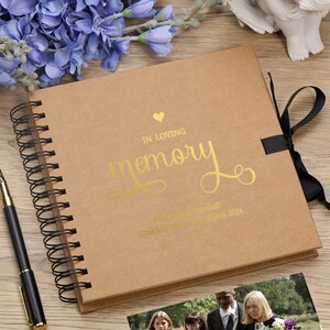 May include: A brown scrapbook with a gold ribbon and a gold inscription that reads "In loving memory" with a heart symbol. The inscription also includes a name and dates.