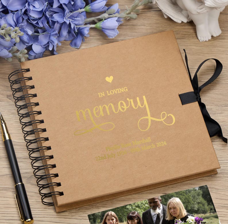 May include: A brown scrapbook with a gold ribbon and a gold inscription that reads "In loving memory" with a heart symbol. The inscription also includes a name and dates.