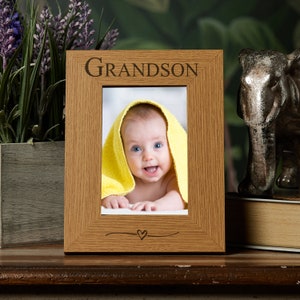 May include: A wooden photo frame with the word "Grandson" engraved on the top. The frame has a small heart engraved at the bottom and a photo of a baby in a yellow towel inside.