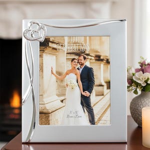 May include: Silver photo frame with a wedding photograph. The frame features a decorative heart design with rhinestones. The photograph is of a couple, and the frame is labelled "Celebrations". The frame is 20cm x 25cm.