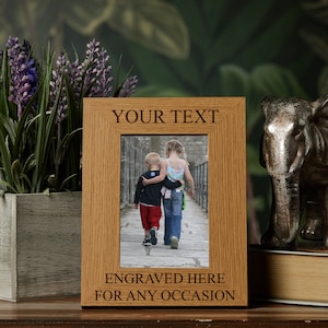 May include: A wooden picture frame with a light brown finish. The frame has a space for a photo and a space for personalised text. The text in the image reads "YOUR TEXT" and "ENGRAVED HERE FOR ANY OCCASION".