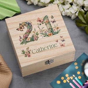 May include: A light-coloured wooden box featuring the number "21" and the name "Catherine" on the lid. The numbers and name are adorned with floral and butterfly motifs. The box is secured with a metal clasp.