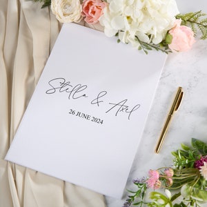 May include: White wedding guest book with the text "Stella & Axel" and the date "26 June 2024" in black script font. A gold pen is lying on a white marble surface next to the book.