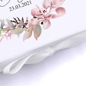 May include: White gift box with a white satin ribbon tied in a bow. The box has a floral design with pink and green watercolor flowers and the date 23.03.2021 printed on the top.
