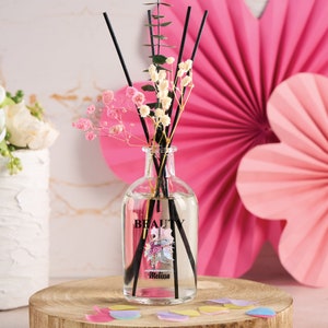 May include: A clear glass bottle diffuser with black reeds and a pink and white floral design. The bottle has a label that says "BEAUTY" and "Melissa".