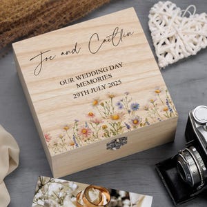 Könnte beinhalten: Helle Holz-Erinnerungsbox mit den Namen "Joe and Caitlin" und dem Datum "29. Juli 2025". Die Box hat einen floralen Rand und den Text "OUR WEDDING DAY MEMORIES". Ein Metallverschluss sichert den Deckel.