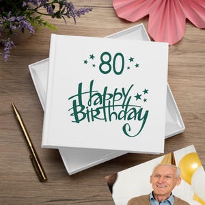 May include: White gift box with a green inscription that reads "80 Happy Birthday" with stars. The box is open and a photo of a smiling man is visible.