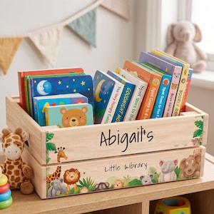 May include: A wooden crate filled with children's books, featuring a variety of colorful covers. The crate is personalized with the name "Abigail's" and "Little Library" on the front, along with jungle animal illustrations. A giraffe and a plush toy are visible.