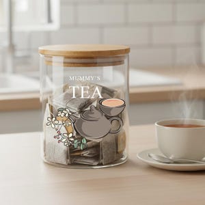 Könnte beinhalten: Ein klares Teeglas mit Holzdeckel, gefüllt mit Teebeuteln. Das Glas hat einen Aufkleber mit dem Text "Mummy's TEA" und einer Teekannen-Illustration. Eine Tasse Tee steht auf einer Untertasse.