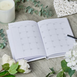 Puede incluir: Una agenda blanca con un dise&ntilde;o de calendario mensual. La agenda est&aacute; abierta en el mes de febrero y el a&ntilde;o 2023. La agenda est&aacute; sobre una mesa de madera con flores blancas y vegetaci&oacute;n.