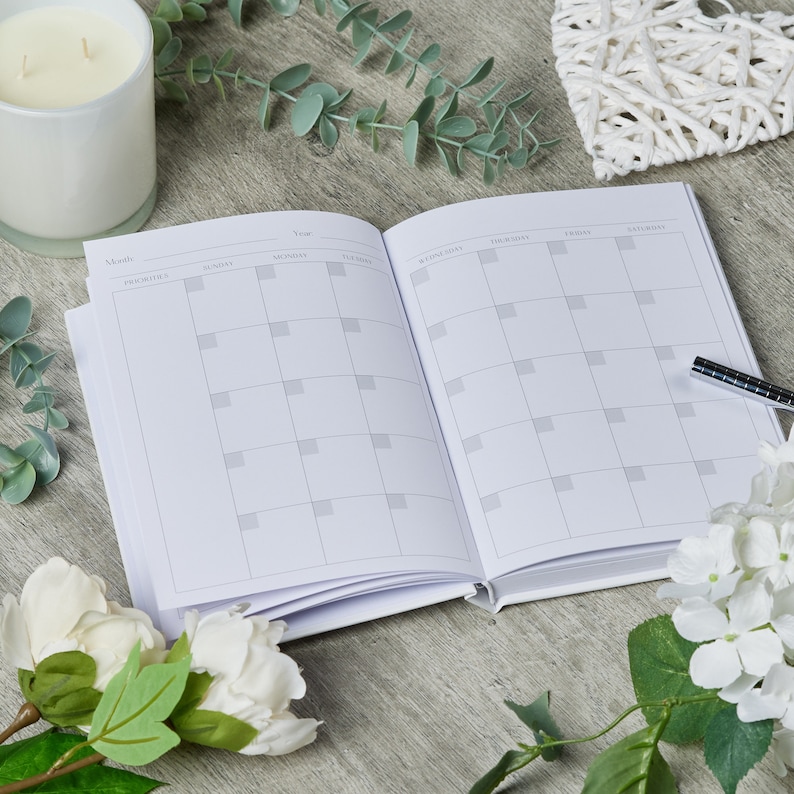 Puede incluir: Una agenda blanca con un dise&ntilde;o de calendario mensual. La agenda est&aacute; abierta en el mes de febrero y el a&ntilde;o 2023. La agenda est&aacute; sobre una mesa de madera con flores blancas y vegetaci&oacute;n.