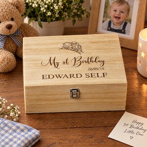 A wooden box with a personalized message engraved on it, placed on a wooden surface next to a teddy bear, a picture frame, and a candle.