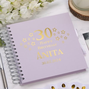 Puede incluir: Un álbum de recuerdos de cumpleaños lila con encuadernación en espiral y letras doradas. La portada dice "30 Feliz Cumpleaños ANITA 30.02.2024" con estrellas. Un bolígrafo plateado es visible a la izquierda.