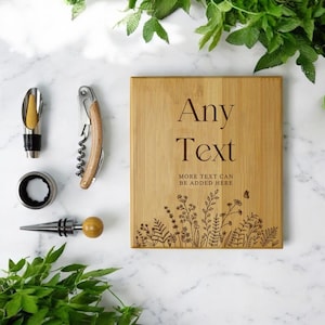 May include: A wooden chopping board with the text "Any Text" and floral engravings, alongside wine accessories: a corkscrew, bottle stopper, and wine pourer. The items are arranged on a white marble surface.