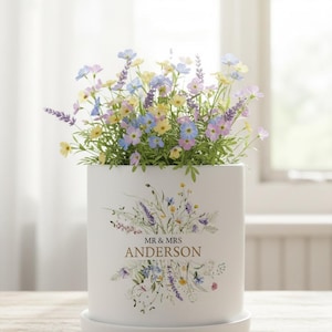 May include: A white ceramic planter, decorated with a floral design and the text "MR & MRS ANDERSON", holds a mix of artificial flowers in yellow, blue, and purple. The planter rests on a matching saucer, a decorative item.