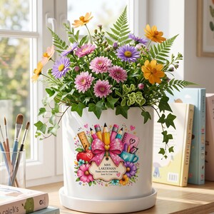 May include: A white ceramic planter with a saucer containing a vibrant floral arrangement of pink, purple, and orange flowers with green ferns. The planter features a colourful graphic of school supplies, a pink bow, and a label reading MRS LAKERMAN Thank you for everything Josh Yr 5 xx.