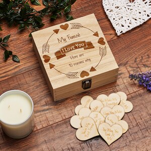 May include: Wooden box with a lid, engraved with the words "My Fianc&eacute; I LOVE YOU Here are 10 reasons why". The box is decorated with hearts and arrows. The box contains small wooden hearts with engraved messages.
