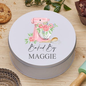 May include: A round, gray metal cookie tin with a pink and white illustration of a stand mixer with flowers. The text "Baked By MAGGIE" is printed on the tin.
