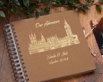 May include: A brown spiral-bound photo album with gold foil lettering and a London skyline design. The cover reads "Our Adventure" and "Natalie & Jack London 2024". A pen rests below the album.